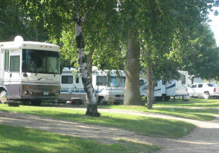 Several RVs and a white pickup truck are parked among green trees in the sunny, grassy Oak Park Campground. A dirt road curves through the scene, with picnic tables visible beside the RVs.