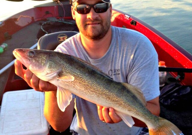 A man wearing sunglasses, a red cap, and a gray t-shirt sits in a red boat holding a large fish. Smiling for the Alexandria fishing report for July, he poses with water in the background and a white cooler beside him.