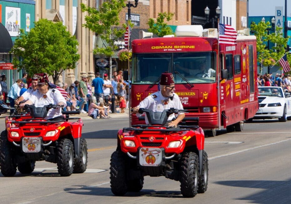 Two Shriners wearing fezzes ride red ATVs in a parade through Alexandria, followed by a red Osman Shrine van. Spectators line the street and American flags are visible in the background, creating a lively Midwest scene.