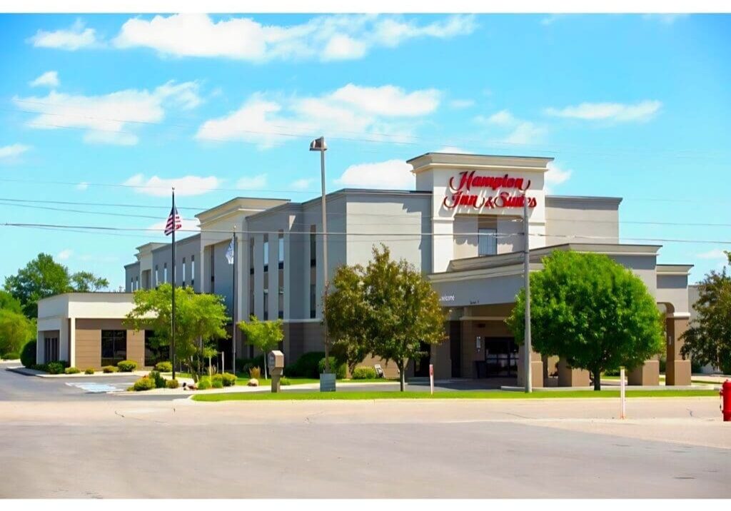 A modern Hampton Inn & Suites hotel building with beige and white exterior, trees, an American flag, and a blue sky with scattered clouds in the background.