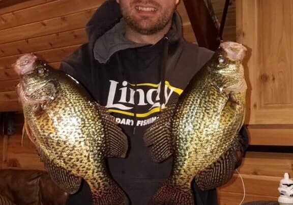 A man in a hoodie and cap smiles while holding two large crappie fish indoors, with wooden walls and fishing gear in the background—showcasing a successful fishing day in Alexandria.