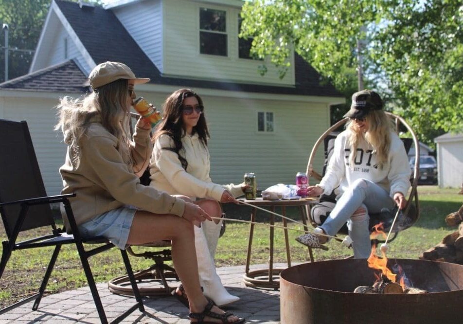 Three women sit around a fire pit in The Howdy House backyard, roasting marshmallows. They wear casual clothes and hats, drinks by their sides. A white house and trees are visible in the background on a sunny day.