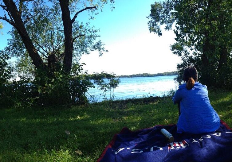 A person in a blue shirt sits on a dark blanket, enjoying a picnic in a park near a calm lake, surrounded by trees and grass with hints of fall color, gazing at the water under a clear, sunny sky.