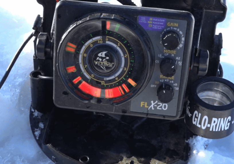 A Vexilar FLX-20 fish finder sits on the snow, displaying colorful sonar readings ideal for winter fishing. Various controls and a Glo-Ring accessory are visible beside the device.