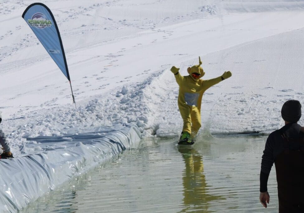 A person in a yellow Teletubby costume snowboards across a pool of water on a snowy slope at Andes Tower Hills during the End of the Season Bash, while a few spectators watch nearby.