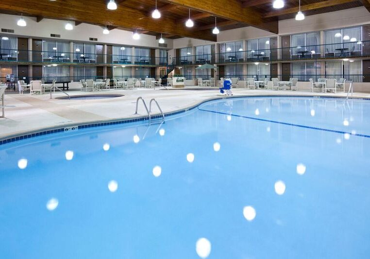 Large indoor swimming pool with clear blue water at Holiday Inn, surrounded by lounge chairs and tables. Overhead lights reflect on the water, while glass-walled hotel rooms line the two-story building around the pool.