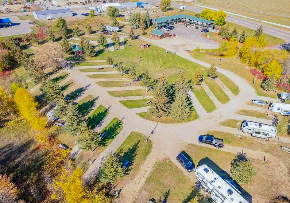 Aerial view of an RV park with several RVs parked on gravel pads among green trees, open grassy areas, and a large building in the background near a road. Some autumn foliage is visible—perfect for your next "RV There Yet" adventure.
