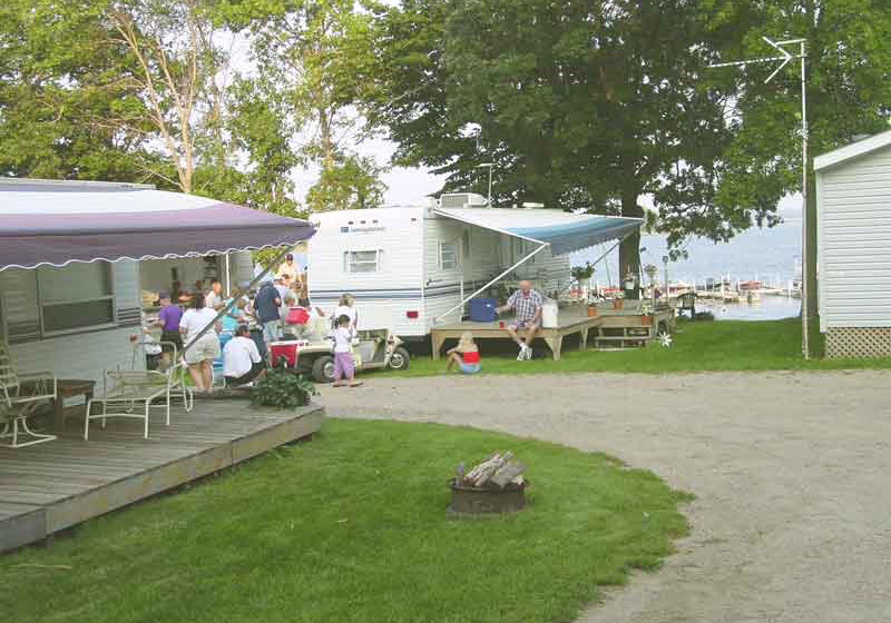 People gather outside RVs at Big Foot Resort Campground, sitting at picnic tables under awnings. Trees, green grass, and a fire pit fill the foreground, while a lake or river shimmers in the background.