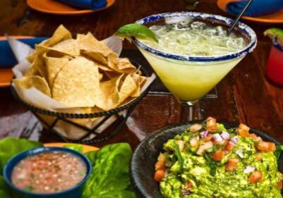 A table at Rancho Viejo with a margarita, a basket of tortilla chips, a bowl of guacamole topped with diced tomato and onion, and a small dish of salsa, all on colorful plates.
