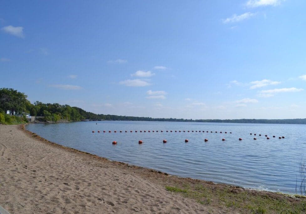 A sandy beach curves along calm lake waters at Rotary Beach Alexandria MN, with a line of red buoys floating under a clear blue sky and green trees surrounding Lake Le Homme Dieu.