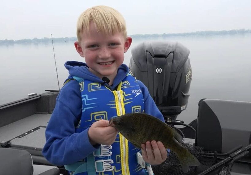 A young boy wearing a blue life jacket smiles while holding a small fish on a boat, enjoying fishing the Midwest with a calm lake and cloudy sky in the background.