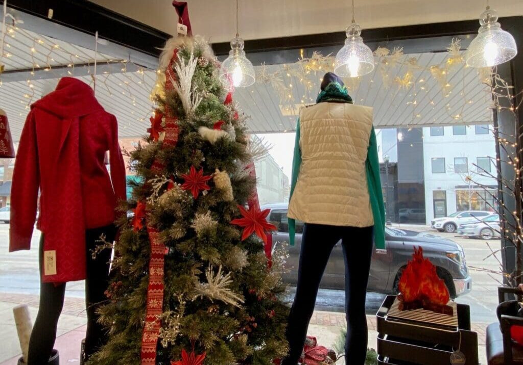 A store window display features two mannequins in winter clothes beside a decorated Christmas tree. Shopping local, you’ll find cozy scenes like this, with a red coat, white vest, faux fireplace, and holiday lights completing the festive atmosphere.