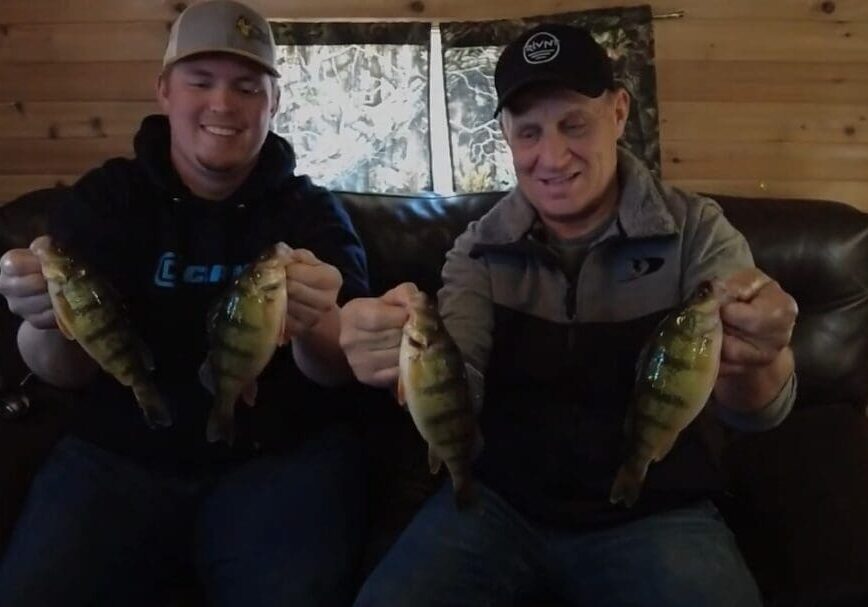 Two smiling men, including Mike Frisch, sit on a couch in a wooden cabin, each holding two fish. The man on the left wears a gray baseball cap and dark hoodie; the man on the right has a black cap and gray jacket.