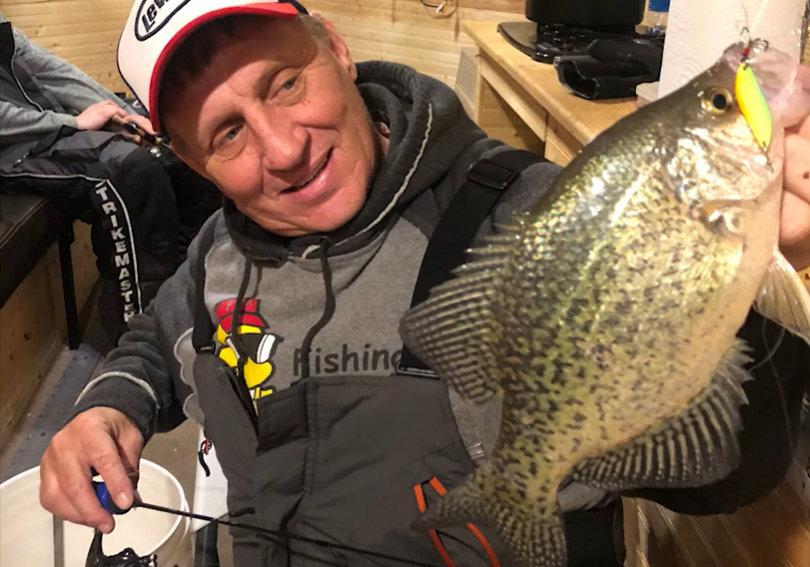 A man in a fishing outfit proudly holds up a large crappie fish with a yellow lure in its mouth, smiling inside a cozy wooden cabin. A fishing rod and bucket highlight the joy of his successful fishing adventure.