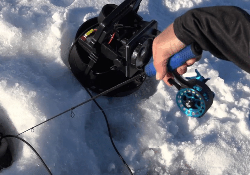 A person holds a blue-handled fishing rod over a hole in the ice, next to a black reel and other ice fishing equipment scattered on the snowy surface.