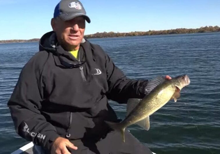 A man in a black jacket and baseball cap sits on a boat during the fishing opener, holding a large fish he caught, with a calm lake and distant tree-lined shore in the background under a clear blue sky.