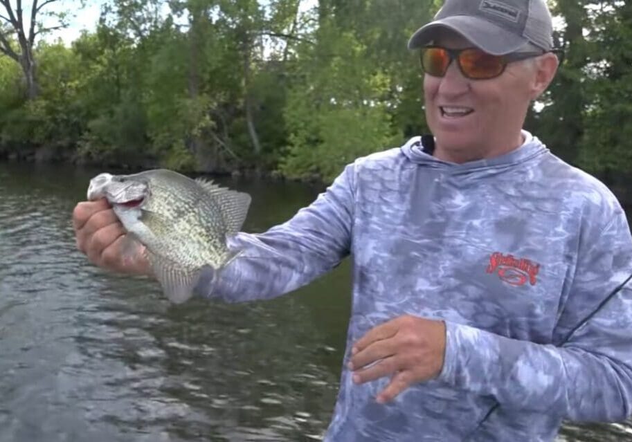 A man wearing sunglasses and a blue camo hoodie holds a large crappie by the water, smiling, with trees in the background—showing off one of the finest crappies of the day.