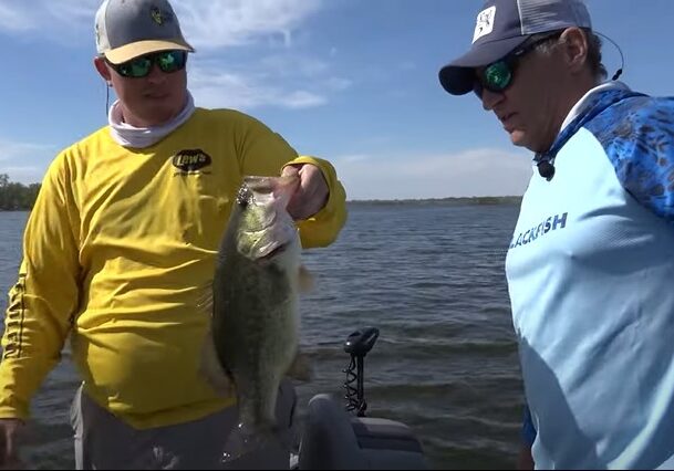 Two men stand on a boat in the water; one, wearing a yellow shirt and hat, proudly holds up a large mouth bass while the other, in a blue shirt and cap, admires the impressive catch. Trees and shoreline are visible in the background.