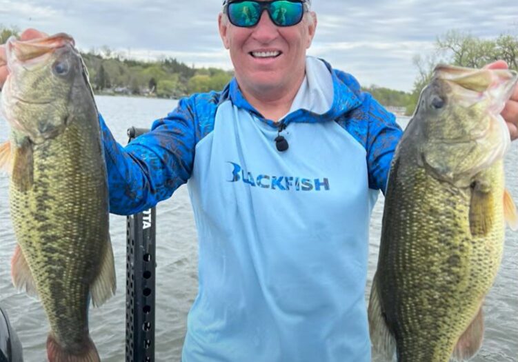 A smiling man in sunglasses and a blue shirt stands on a boat, holding up two large bass fish caught with a squarebill, with water and trees in the background.