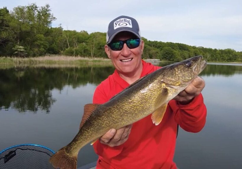 A man in a red shirt and cap smiles while holding a big fish, standing proudly in a boat on a calm lake with trees in the background.