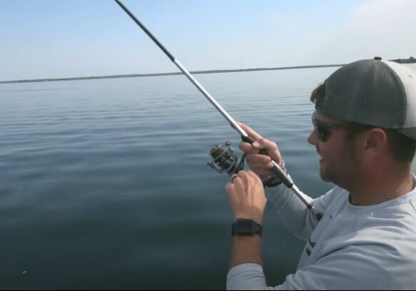 A man wearing a gray cap and sunglasses is holding a fishing rod over calm water, appearing to reel in a catch with swimbaits on a sunny day.
