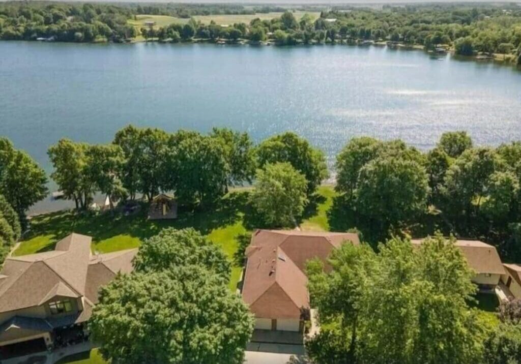 Aerial view of houses and tree-lined yards along the edge of calm Lake Latoka, with open green fields and wooded areas in the background under a clear sky—perfect for a peaceful family retreat.