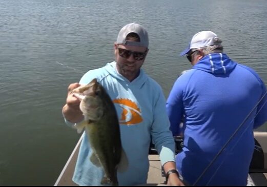 Two people on a boat; one man in sunglasses and a light blue hoodie, proudly holding up a big fish and smiling at the camera, while the other in a blue shirt faces away, fishing on a calm lake.