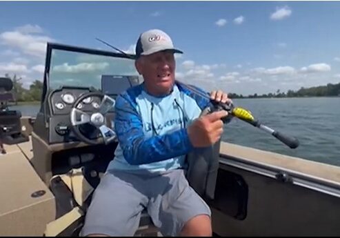 A man in a blue long-sleeve shirt and cap sits in a boat on a lake, holding a fishing rod with moving baits. The sky is clear with scattered clouds, and trees are visible in the background.