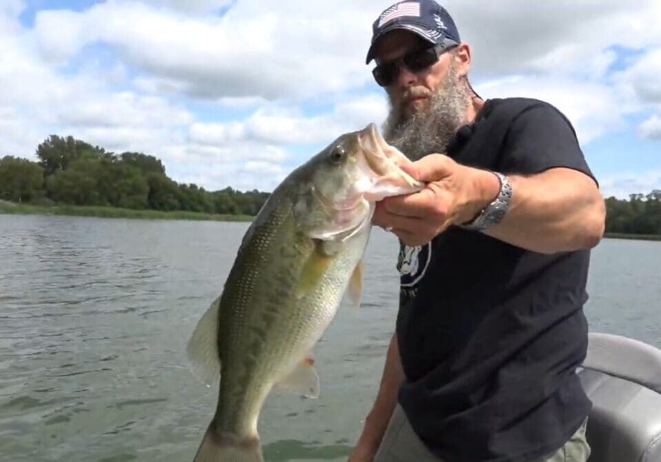 A bearded man wearing sunglasses and a cap holds a large fish on a boat, enjoying some fall fishing, with a lake and trees in the background under a partly cloudy sky.