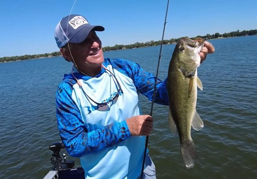 A man in a blue fishing shirt and cap holds up a large fish he caught while fishing the Midwest from his boat on a lake, with clear blue water and a distant tree-lined shore under a sunny sky.