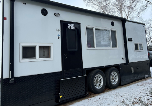 A white and black mobile trailer labeled "MDNR" with two windows, two wheels, and a black door stands on snowy ground by leafless trees—perfect for winter outings like Nathan's 7-Holer Fish House.