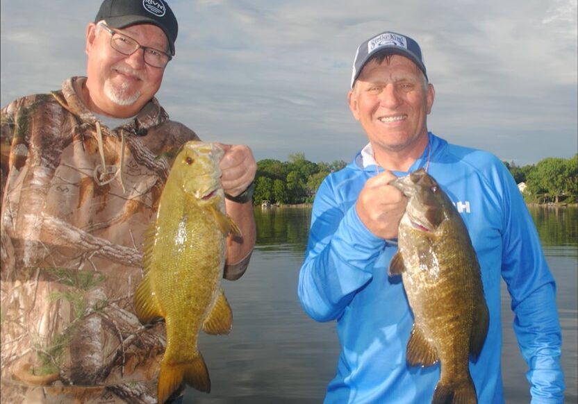 Two smiling men in outdoor clothing and hats proudly hold up large fish while standing near a calm lake—showing off how to catch more summer bass with ease amid trees and houses in the background.