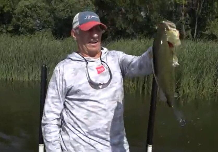 A man in a gray hoodie and red cap holds up a large fish next to a body of water in the Alexandria Lakes Area, with tall grass and trees in the background.