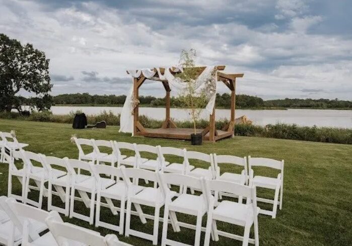 An outdoor wedding ceremony setup at Rendezvous Farm with rows of white chairs facing a wooden arch draped in white fabric, set on green grass near a lake under a cloudy sky.