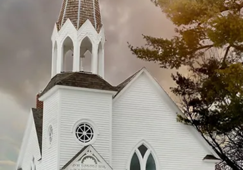 A white wooden Farwell Historic Church with a tall steeple and arched windows stands under a dramatic cloudy sky. A wreath-adorned sign in front reads "Raswell Norwegian Cemetery 1907." Snow covers the ground and trees frame the scene.