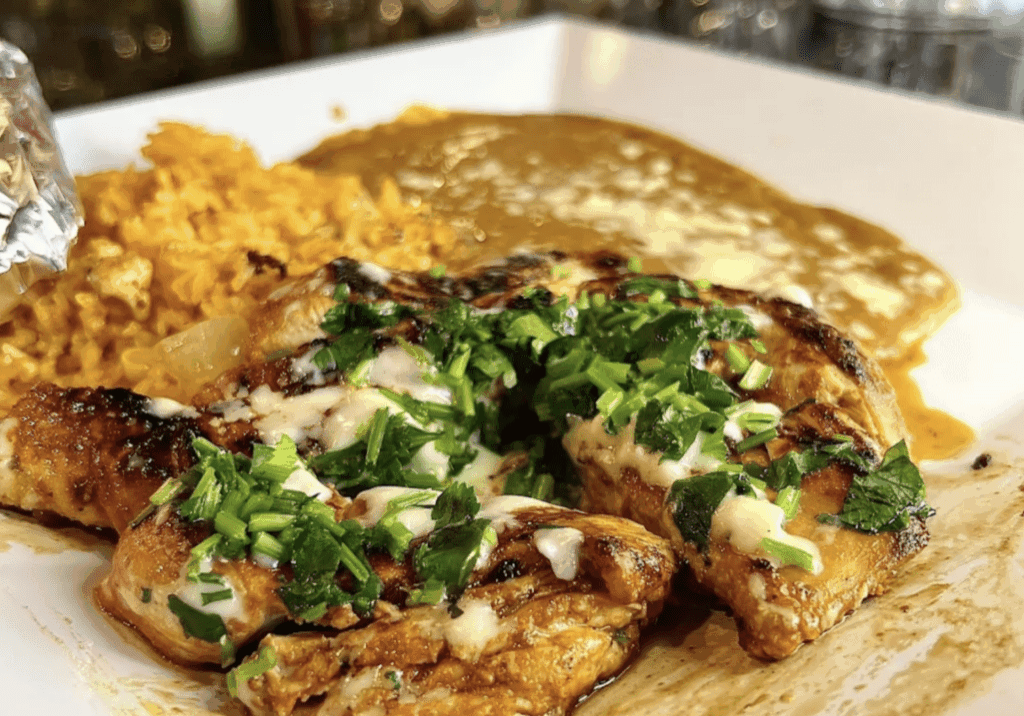 Grilled chicken breast topped with chopped herbs and melted cheese, served with yellow rice and a creamy brown sauce on a white plate—a flavorful El Loro specialty. Glassware is visible in the background.
