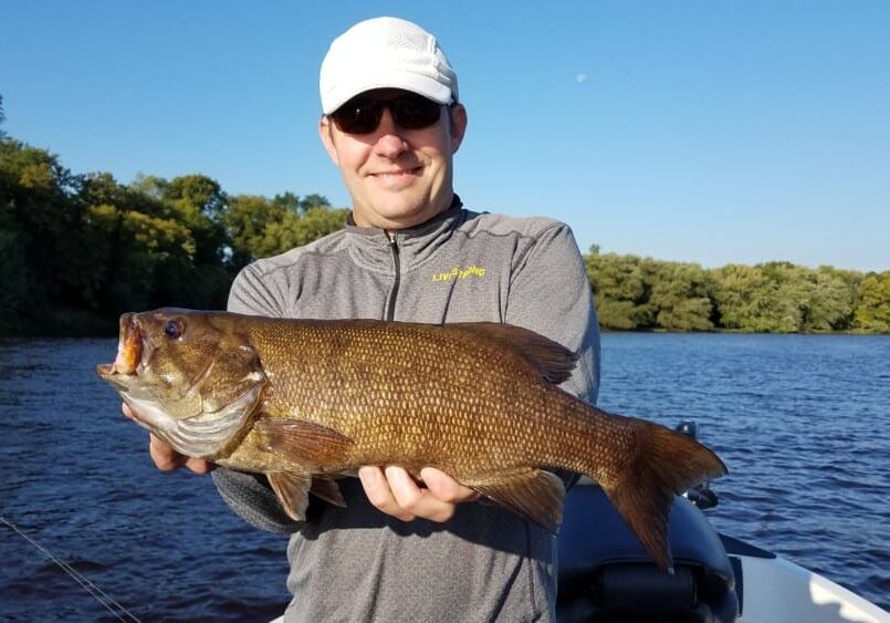 A man wearing sunglasses and a white cap holds a large brown fish while standing on a boat in the lake, enjoying the aggressive fall bite in the Alexandria Lakes area, with trees and a clear blue sky in the background.