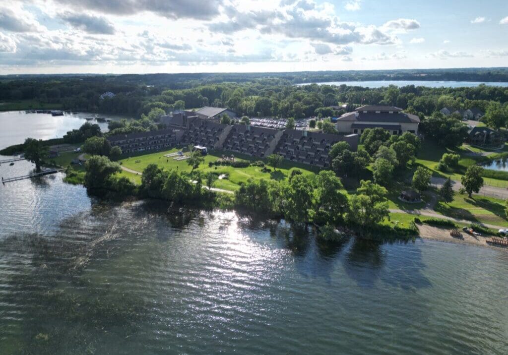 Aerial view of Arrowwood Resort, a large building complex surrounded by trees, located next to a lake with calm water. The sky is partly cloudy, and sunlight reflects off the water’s surface.