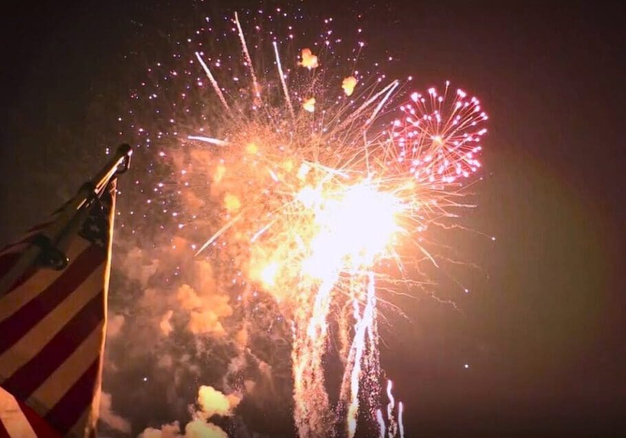 A brightly lit night sky filled with colorful fireworks. In the foreground, part of an American flag is visible, suggesting a festive or patriotic 4th of July celebration in the lakes area.