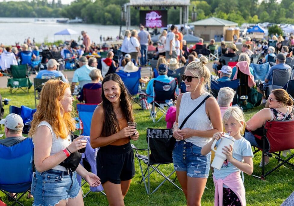 A crowd enjoys Brophy’s 2024 Summer Sounds by a lake. Three women and a girl stand and smile in the foreground, while people sit in lawn chairs facing a large screen. It’s a sunny day filled with music and laughter.