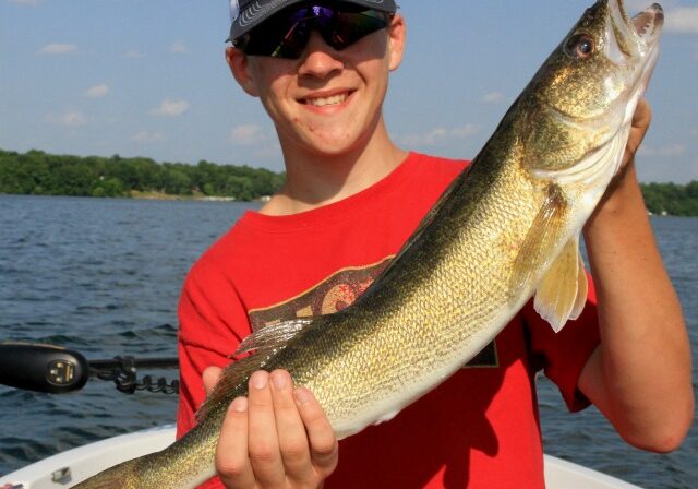 A smiling person in a red shirt and cap holds a large fish on a boat in a lake, showcasing summer fishing techniques against a backdrop of trees and blue sky.