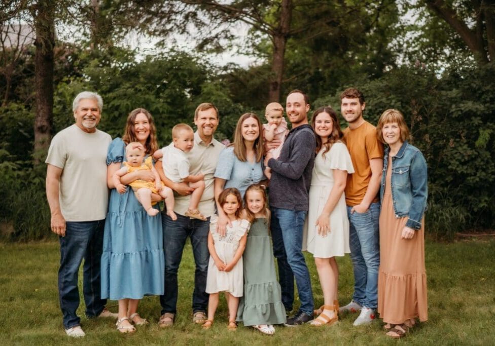 A large family poses together outdoors, smiling. Cindy Swenson captures six adults, three small children, two young girls, and a baby in casual summer clothes standing on grass with trees and bushes in the background.