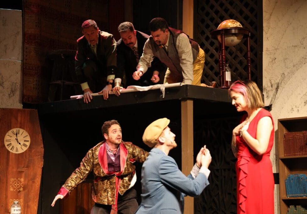 Five men in vintage costumes act dramatically on a two-level stage, with three leaning over a balcony above. Below, one man kneels, holding a ring to a surprised woman in red—an engaging proposal scene for your weekend theatre plans.