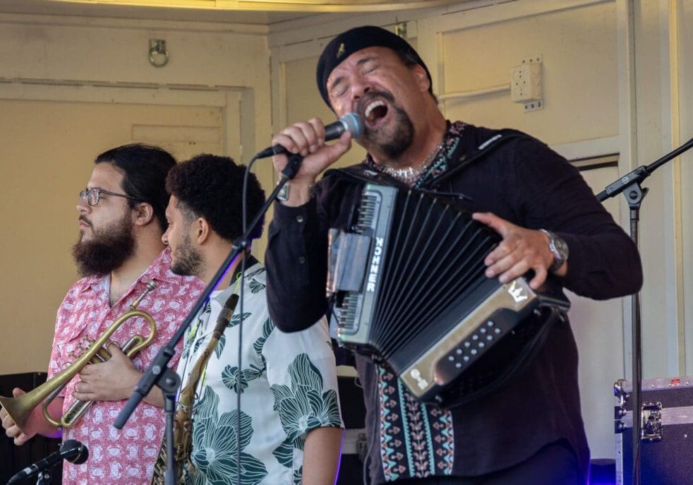 Three musicians perform on stage in Alexandria. One sings passionately into a microphone while playing an accordion, evoking a true Zydeco Experience, while the other two, holding brass instruments, look to the side. All wear patterned shirts.