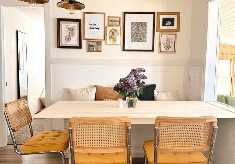 The Darling Cabin’s dining area features a light wooden table, three yellow-cushioned chairs, wall art above, a vase of purple flowers, and pendant lights on a white paneled ceiling. Natural light pours in from the window.