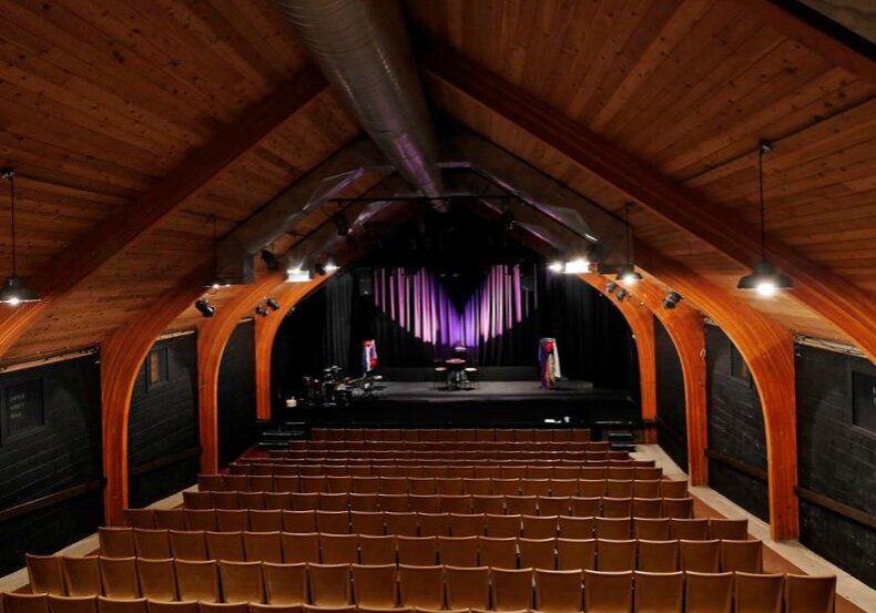 A view of an empty Theatre L’Homme Dieu, showcasing wooden arched ceilings, rows of brown seats, and a stage featuring vertical light bars and two music stands with colorful cloths draped over them.