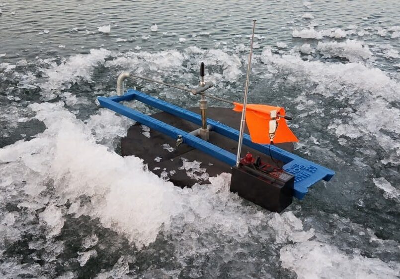A small blue and black robotic device with an orange flag floats among chunks of ice on the surface of a body of water in the Alexandria area.