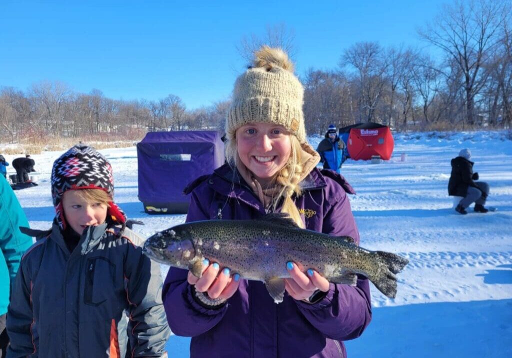 A smiling woman in a knit hat and purple coat holds a large fish on a snowy, frozen lake during an ice fishing trip. A boy stands beside her, with people ice fishing in shelters visible in the background under a clear blue sky.