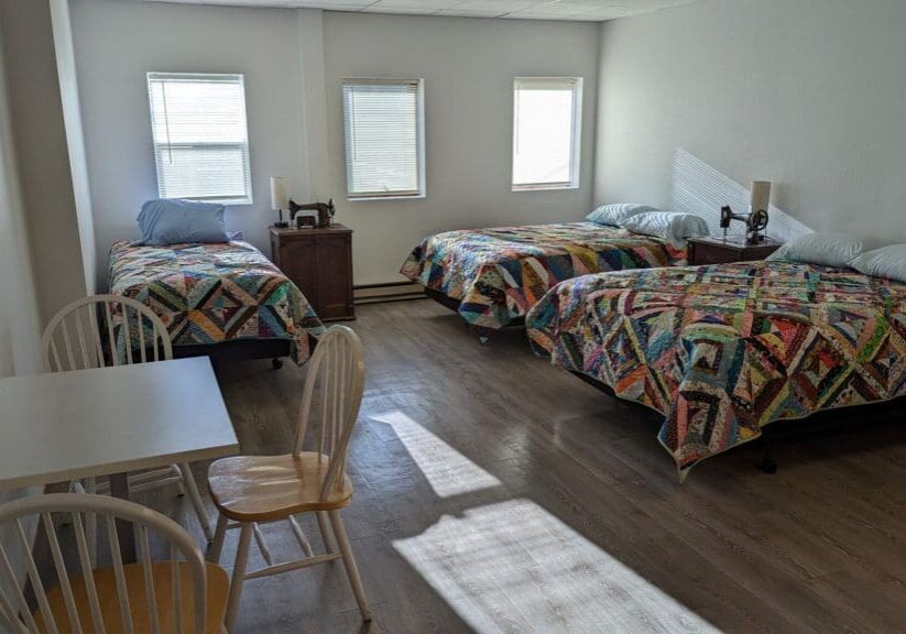 A bright room at Dawn's Quilt Shop & Retreat Center features three beds with colorful patchwork quilts, two nightstands with lamps, a small desk with two chairs, and sunlight streaming through three windows.