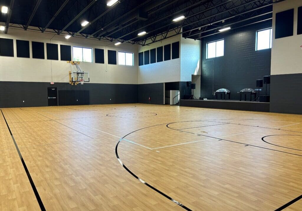 An empty indoor basketball court with a wooden floor, black lines, and high windows. There is a basketball hoop at one end, a stage area with speakers and tables on the right, and room for pickleball games—part of the versatile VIE Church gym.